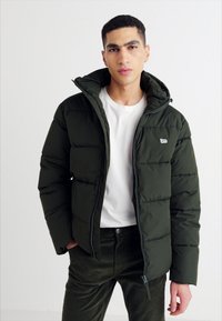 Giacca a vento verde scuro con zip frontale, cappuccio e design trapuntato. Indossata sopra una t-shirt bianca, abbinata a pantaloni scuri.