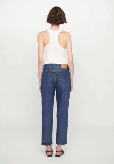 Top sin mangas blanco, jeans de tiro alto azul con patas rectas y bolsillos traseros visibles. Etiqueta de cuero en la cinturilla. Sandalias planas.