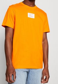 Hombre con una camiseta de Calvin Klein Jeans de color naranja brillante con un pequeño logo blanco en el pecho, combinada con jeans de denim azul claro.