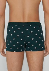 Mann in dunkelgrünen Boxerbriefs, die mit verstreuten schwarzen 8-Ball-Billard-Motiven bedruckt sind, von hinten gezeigt.