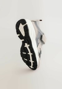Szare i białe sneakersy z czarną teksturowaną podeszwą, górą z siateczki i zamszu oraz z paskiem na rzep, ustawione pod kątem, pokazujące wzór podeszwy.