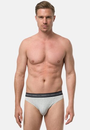 Muskuløs mand stående med front mod kameraet, iført lysegrå underbukser med en mørk linning mærket "nur der" mod en ensfarvet baggrund.