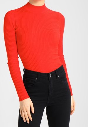 Femme portant un pull col roulé rouge côtelé à manches longues et ajusté, ainsi qu'un jean noir taille haute, debout les mains détendues.