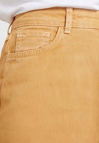 Pantalones de algodón color beige con una superficie texturizada, bolsillo lateral visible y costuras detalladas. Sin elementos metálicos o adornos visibles.