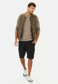 Leichtgewichtige, olivgrüne gesteppte Weste über einem beigen Langarmshirt und marineblauen Cargo-Shorts, kombiniert mit weißen und schwarzen Sneakers.