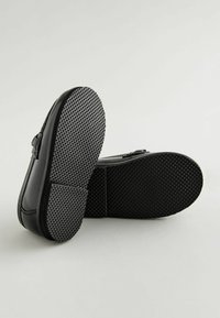 Zapatos negros tipo slip-on con un acabado brillante, que cuentan con una suela de goma texturizada y una hebilla decorativa en el lateral. Las suelas están diseñadas con un patrón para mejorar el agarre.