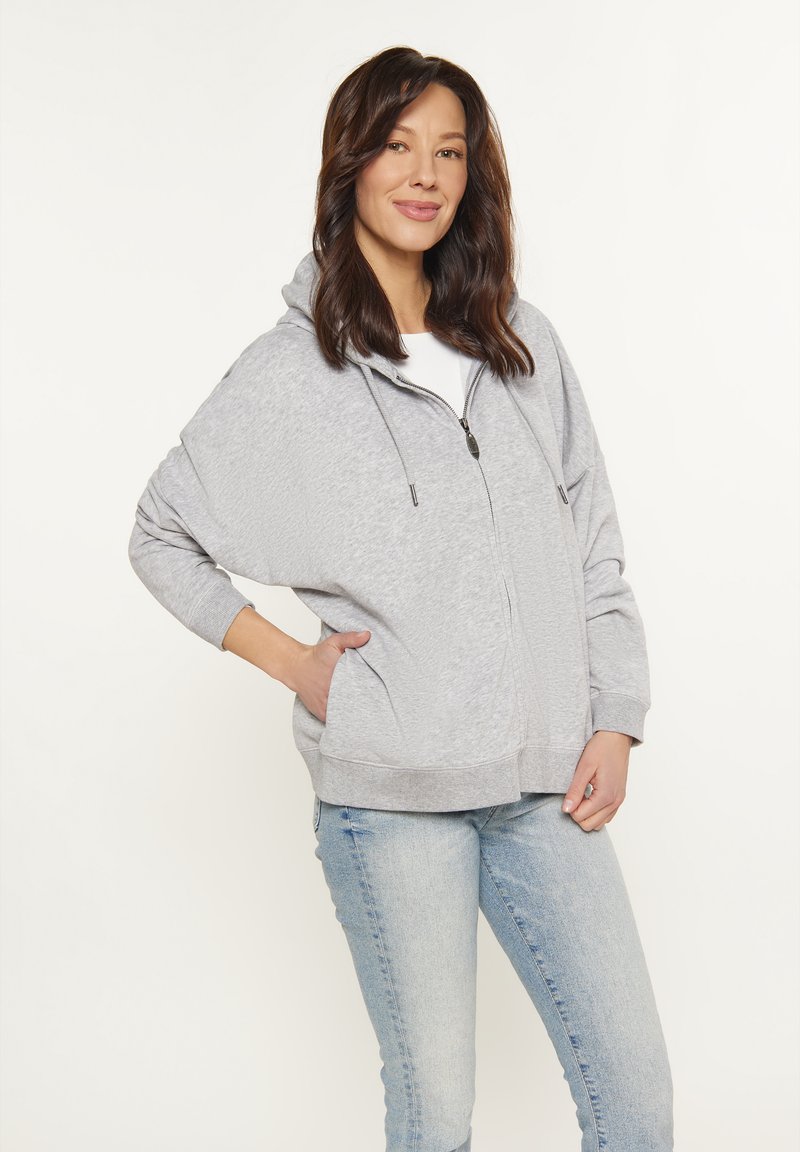 Felpa grigia chiara con zip, dal taglio morbido, grandi tasche e cappuccio regolabile. Indossata sopra una maglietta bianca, abbinata a jeans blu chiaro.