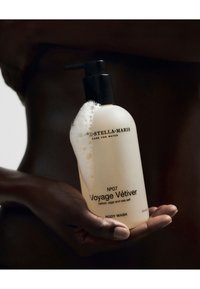 Bouteille en plastique transparente avec une pompe noire, contenant un gel douche mousseux blanc étiqueté "Voyage Vétiver" en texte noir. Fini mat doux.