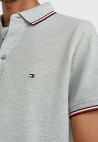 Helle graue Polo-Shirt mit einem Kragen und Ärmelabschlüssen, die mit schwarzen, weißen und roten Streifen versehen sind, sowie einem kleinen rechteckigen Logo auf der Brust.