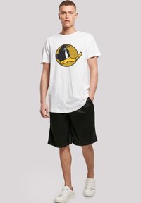Hvit bomulls t-skjorte med et gult og svart sirkulært grafisk motiv av en tegneserie-and, kombinert med svarte shorts og hvite joggesko.