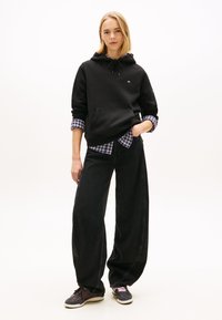 Νεαρή γυναίκα που στέκεται φορόντας μαύρο φούτερ με κουκούλα πάνω από μπλε πουκάμισο με καρό, φαρδιά μαύρα τζιν και σκούρα αθλητικά παπούτσια με παστέλ κορδόνια.