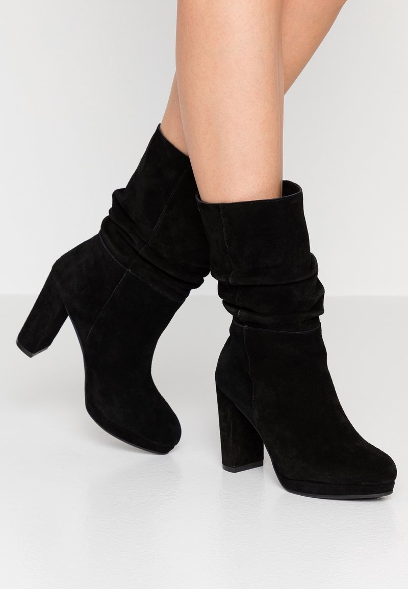 Black suede ankle boots with a slouchy design, high block heel, and rounded toe. Soft texture visible, with minimal stitching detail.