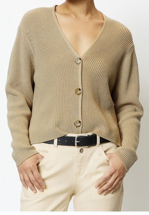 Personne portant un cardigan en maille beige boutonné sur un pantalon crème avec une ceinture noire, les mains dans les poches.