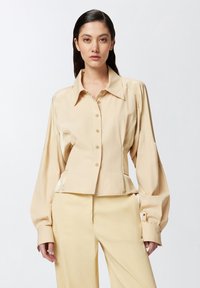 Bež košulja skraćena do struka s strukturiranim ovratnikom, dugim rukavima i prednjim kopčanjem na dugmiće. Izrađena od glatkog materijala s suptilnim sjajem.