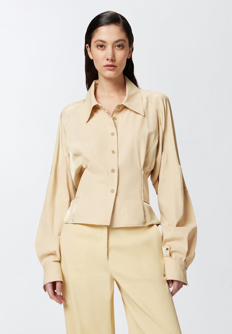 Bež košulja skraćena do struka s strukturiranim ovratnikom, dugim rukavima i prednjim kopčanjem na dugmiće. Izrađena od glatkog materijala s suptilnim sjajem.