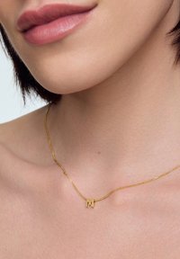 Zbliżenie na szyję i usta osoby noszącej cienki złoty naszyjnik z małym pendantem w kształcie litery "M".