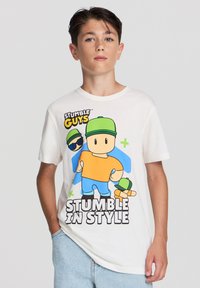 Biała koszulka z krótkim rękawem z kolorową grafiką przedstawiającą postacie w zielonych czapkach oraz napisem "STUMBLE IN STYLE." Luźny krój.