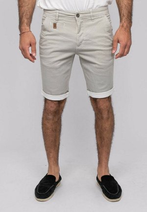 Homme portant un short beige texturé jusqu'aux genoux avec des revers, une chemise blanche rentrée et des chaussures noires sans lacets sur un fond uni.