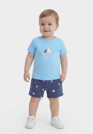 Bambino piccolo che indossa una maglietta azzurra con grafica di una barca a vela, pantaloncini blu navy con motivi di barche a vela e scarpe da ginnastica bianche, in piedi e sorridente.