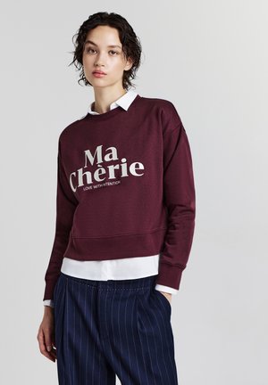 Junge Frau mit kurzen lockigen Haaren, die einen burgunderroten Pullover mit der Aufschrift "Ma Chérie" über einem weißen Hemd und einer navyblauen Nadelstreifenhose trägt.