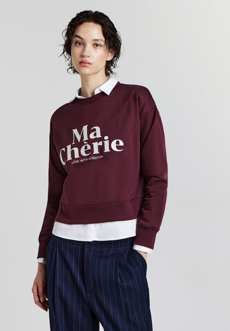 Giovane donna con capelli ricci corti che indossa una felpa bordeaux con la scritta "Ma Chérie" sopra una camicia bianca e pantaloni a righe blu navy.