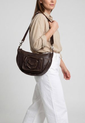 Frau trägt beige Bluse und weiße Hose und trägt eine dunkelbraune Lederschultertasche mit vorderen Taschen und Reißverschlüssen.