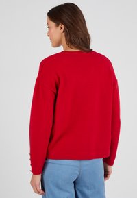 Maglione rosso lavorato a maglia con una vestibilità rilassata, caratterizzato da spalle scese e bottoni laterali sulle maniche. Texture morbida e orlo a coste.