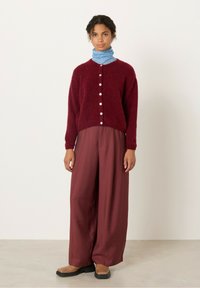 Bordo pleteni kardigan z belimi gumbi, položen preko modrega črtastega puloverja z visokim ovratnikom. Široke, lahkotne bordo hlače in rjave čevlje dopolnjujejo videz.