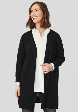 Cardigan noir à ouverture frontale, à manches longues et à ourlet droit. Porté par-dessus une chemise blanche à boutons, avec un tissu doux et une coupe décontractée.