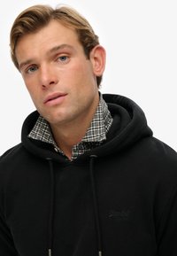 Sudadera negra con cordones sobre un cuello de camisa a cuadros en verde y blanco. La sudadera presenta un logotipo sutil y una textura suave.