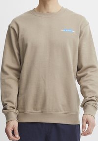 Sweat-shirt beige avec poignets et ourlet côtelés, encolure ronde, et un petit motif bleu indiquant "TOKYO" sur la poitrine. Texture de tissu douce.