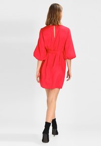 Červené krátké šaty s páskem v pase, nafouknutými rukávy a detailem ve tvaru klíčové dírky na zádech, doplněné černými kotníkovými botami. Hladká textura.