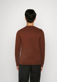Sudadera larga marrón de manga larga con cuello redondo, hecha de material suave, con un dobladillo y puños de canalé, mostrada desde atrás.