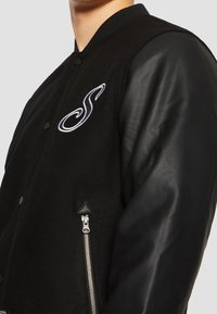 Zwarte bomberjack met een wollen lichaam, leren mouwen, drukknoopsluiting, geborduurd "S"-logo en een ritszak met een zilveren trekker.
