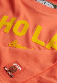 T-shirt corail avec un texte "HOLA" en jaune audacieux, présentant une finition texturée. Inclut une petite étiquette de marque avec un détail de logo en relief.