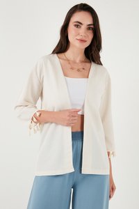 Mujer con cárdigan beige abierto al frente con flecos en las mangas, top corto blanco, pantalones anchos azules y collares dorados en capas.
