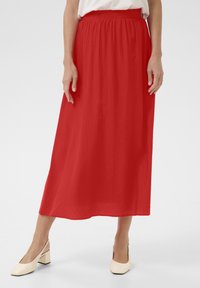 Jupe rouge de mi-longueur en tissu léger avec une taille élastique. Texture lisse, silhouette fluide. Assortie à des chaussures à talons carrés beiges.