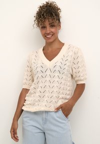 Pulover crem tricotat cu decolteu în V și mâneci scurte tip puff, prezentând un model cu frunze; asortat cu pantaloni cu dungi albastre deschise.