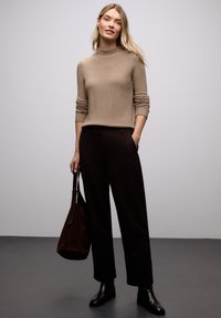 Pull en maille côtelée beige à col rond, associé à un pantalon noir large taille haute. Le modèle tient un sac en daim marron foncé et porte des bottines.