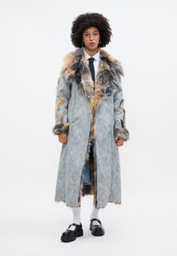 Garšs pelēks mētelis ar mākslīgās kažokādas apdari oranžā un melnā krāsā. Tam ir sānu izgriezumi un teksturēts apdare. Valkāts virs džinsiem un saskaņots ar melnām kurpēm.