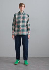 Grønn og beige rutete skjorte med knapper, kombinert med mørkeblå bukser og lysegrønne joggesko. Plassert mot en nøytral bakgrunn.