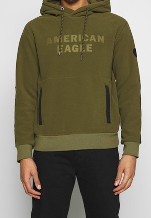 Muž nosící olivově zelenou mikinu s kapucí s nápisem "AMERICAN EAGLE", šňůrkami, kapsami na zip vpředu a černými kalhotami na jednobarevném pozadí.