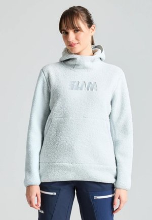 Una mujer luce una sudadera blanca de forro polar con capucha y bolsillo delantero, y unos pantalones azul marino, sobre un fondo gris liso.