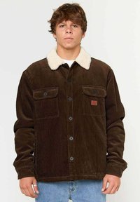 Brun fløjljakke med en cremefarvet faux fur krave, to brystlommer og sorte knaplukninger, båret over en hvid skjorte.