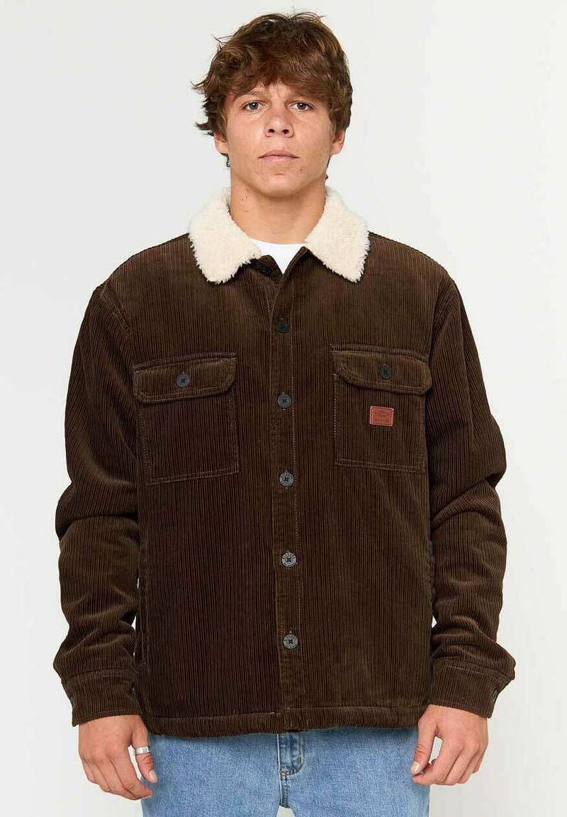 Brun fløjljakke med en cremefarvet faux fur krave, to brystlommer og sorte knaplukninger, båret over en hvid skjorte.