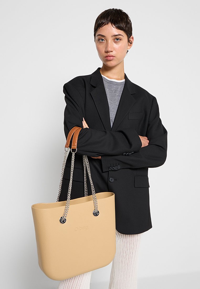 Beżowa torba tote o gładkiej teksturze, z łańcuchami w kolorze srebrnym oraz brązowymi skórzanymi akcentami. Torba ma szeroki, otwarty design.