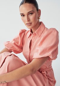 Ružičasta haljina s gumbima, napuhanim rukavima i vezanim pojasom. Tekstil izgleda lagano s glatkom teksturom i jednostavnim dizajnom.