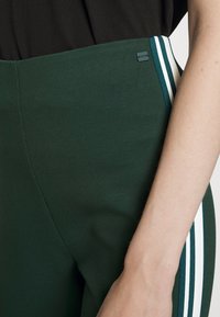 Gröna högmidjade byxor med strukturerad tyg. Har vita sidostripes och en liten varumärkesetikett nära midjan.