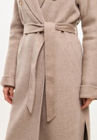 Cappotto in misto lana beige con design avvolgente, dotato di cintura e spacchi laterali. Dettagli notevoli includono bottoni grandi e tessuto testurizzato.