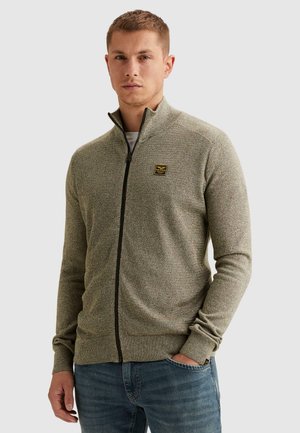 Joven con un suéter de punto beige con cremallera y jeans azules, con una mano en el bolsillo, expresión neutral, fondo liso y claro.